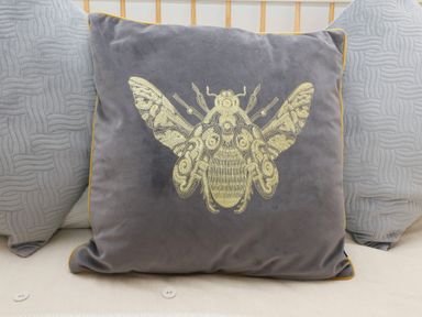 A gray decorative cushion with a gold embroidered insect design on a beige surface with two gray cushions behind at Bowderstone in Keswick