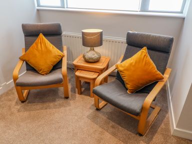 Two wooden armchairs with gray cushions and orange pillows beside a wooden side table with a lamp in a carpeted corner room at Bowderstone in Keswick