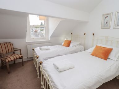 A bedroom with two single beds with white bedding and orange cushions a striped armchair and a window at Number Twenty in Keswick