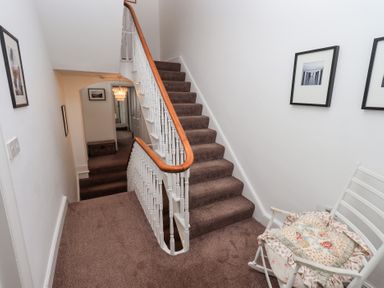 A staircase with brown carpet and white railing a white chair with a cushion and framed pictures on the wall at Number Twenty in Keswick