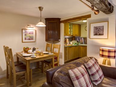 A dining area with a wooden table and chairs set for breakfast next to a kitchen with wooden cabinets and a living area with a sofa and plaid cushions at Burrow Cottage in Bowness