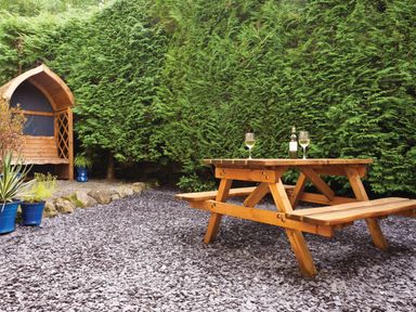 An outdoor seating area with a wooden picnic table and bench with two glasses and a bottle on the table next to a wooden arched garden bench and potted plants