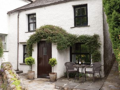 A cottage entrance with a wooden door and two potted plants beside it and a small table with two chairs and wine glasses at Burrow Cottage in Bowness