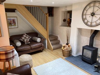 A living room with a leather sofa under stairs a fireplace with firewood and a large clock on the wall at Burrow Cottage in Bowness