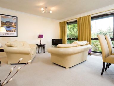 A living room with cream sofas a black TV a purple lamp a dining table with chairs and large windows with yellow curtains at Mount Sol in Ambleside