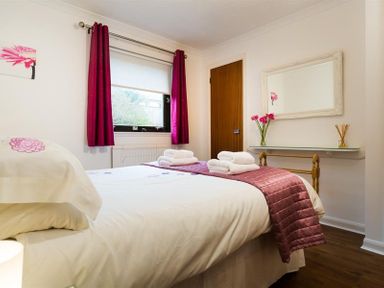 A bedroom with a bed with white linens and pink accents a window with pink curtains a wooden door and a wall-mounted shelf with a mirror and flowers at Mount Sol in Ambleside