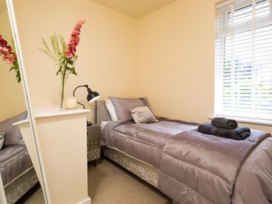 A bedroom with a single bed covered with grey bedding beside a window with blinds and a bedside table with a lamp and flowers at Greengarth in Keswick