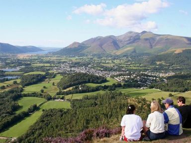Four people sitting on a hill overlooking a valley with fields trees a town and mountains in Keswick