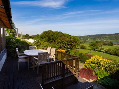 An outdoor wooden deck with a table and chairs overlooking a green field and distant hills at Garden Cottage At Coniston in Coniston