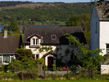 A white cottage with a dark roof surrounded by greenery and flowers with a body of water and forested hills in the background at Garden Cottage At Coniston