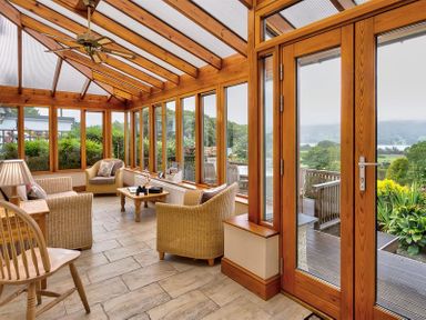 A sunroom with wicker chairs, a wooden table, a wooden chair, and glass doors overlooking a garden at Garden Cottage At Coniston