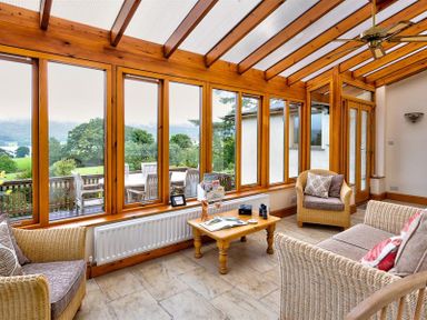 A sunroom with wicker chairs a wooden coffee table and large windows overlooking a garden at Garden Cottage At Coniston in Coniston
