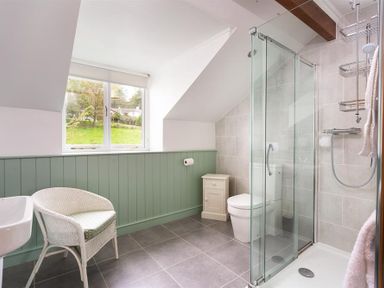 A bathroom with a glass shower, white toilet, small cabinet, wicker chair, and window at Garden Cottage At Coniston in Coniston