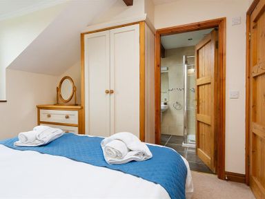 A bedroom with a bed covered by a blue quilt and white towels on it a white wardrobe and a wooden dresser with a round mirror a wooden door leading to a bathroom at Garden Cottage At Coniston in Coniston