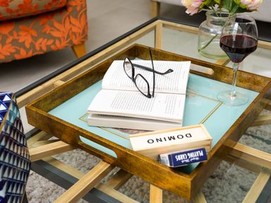 A glass coffee table with a wooden tray holding an open book with glasses a domino set a deck of playing cards and a glass of red wine near an orange patterned chair