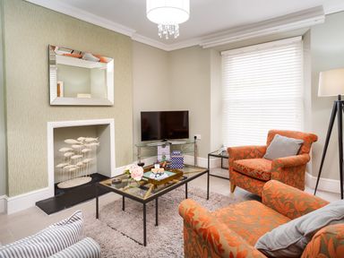 A living room with orange patterned armchairs a glass coffee table a television a mirror above a fireplace and white blinds at Pippin Cottage in Keswick
