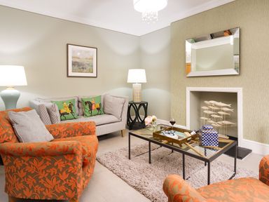 A living room with orange patterned armchairs a grey sofa with green cushions a glass coffee table and table lamps at Pippin Cottage in Keswick