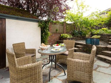 An outdoor patio with a glass table surrounded by four wicker chairs next to a garden and stone steps at Pippin Cottage in Keswick
