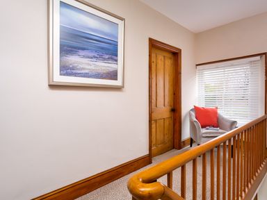 A hallway with a wooden door a framed painting a gray chair with a red cushion near a window with blinds and a wooden railing at Pippin Cottage in Keswick