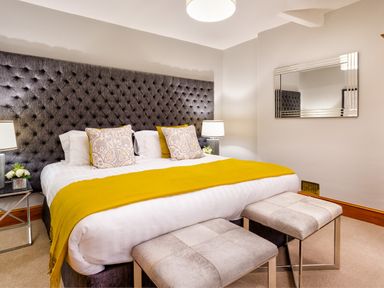 A bedroom with a large bed featuring a tufted headboard two cushioned stools at the foot a yellow throw and patterned pillows at Pippin Cottage in Keswick