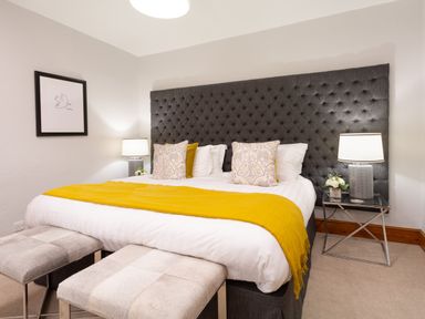 A bedroom with a large bed with a grey tufted headboard yellow blanket and two benches at Pippin Cottage in Keswick