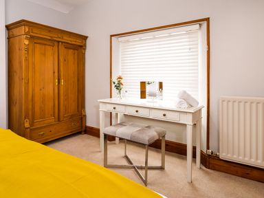 A bedroom with a wooden wardrobe a white dressing table a bench and a bed with yellow bedding at Pippin Cottage in Keswick