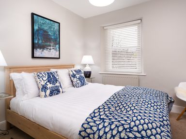 A bedroom with a wooden bed white bedding blue patterned pillows and a matching blanket a window with blinds a lamp on a side table and a painting on the wall at Pippin Cottage in Keswick