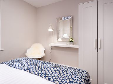 A bedroom with a bed covered by a blue patterned blanket a white chair with a sheepskin throw a wall shelf with a mirror lamp and flowers and a white wardrobe at Pippin Cottage in Keswick