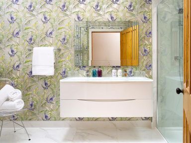 A bathroom with a wall-mounted white sink cabinet a mirror patterned wallpaper and a glass shower enclosure at Pippin Cottage in Keswick