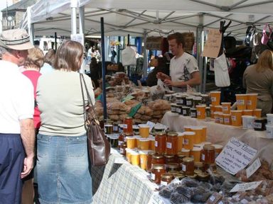 An outdoor market stall with jars of preserves and baked goods with people browsing at a market