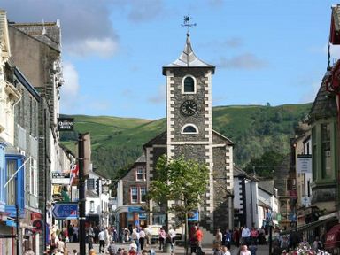 A busy street with a clock tower in the center and shops on both sides with hills in the background at Pippin Cottage in Keswick