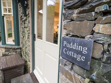 An exterior view of a stone cottage wall with a sign reading Pudding Cottage and a white door at Pudding Cottage in Ambleside