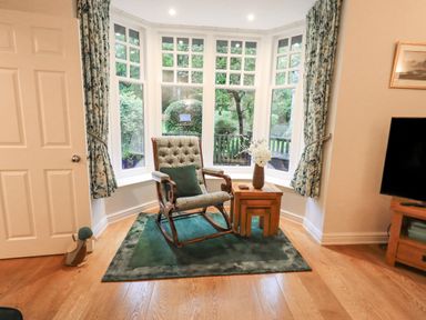 A sitting area with a rocking chair a small wooden table with a vase and flowers in front of bay windows with patterned curtains at Pudding Cottage Ambleside