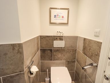 A small bathroom with a toilet paper holder a toilet brush a framed picture on the wall and large rectangular tiles on the walls at Pudding Cottage in Ambleside