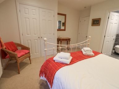 A bedroom with a double bed with towels on a red throw a wicker chair a wooden side table mirror closet doors and an open door to a bathroom at Pudding Cottage in Ambleside