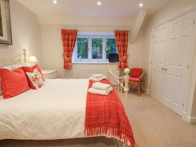 A bedroom with a white bed and red cushions and blanket near a window with red curtains and a wicker chair at Pudding Cottage in Ambleside