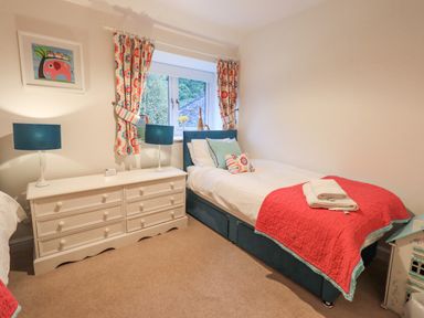 A bedroom with a single bed beside a white dresser with two blue lamps and a window with patterned curtains at Pudding Cottage in Ambleside