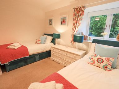 A bedroom with two single beds a white dresser with lamps and a window with floral curtains at Pudding Cottage in Ambleside
