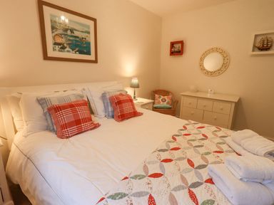 A bedroom with a bed with red and blue checked pillows a white dresser a wicker chair and framed pictures at Pudding Cottage Ambleside