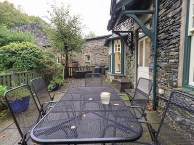 An outdoor patio with a black metal table and six chairs next to a stone cottage with a white door and green trim at Pudding Cottage Ambleside