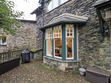 Stone exterior wall with a green framed bay window overlooking a gravel area with a covered barbecue and storage boxes at Pudding Cottage in Ambleside