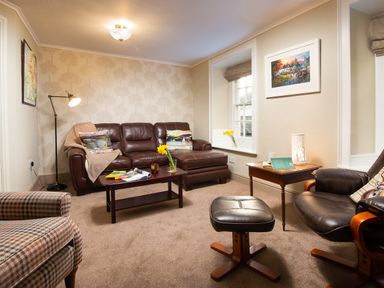 A living room with leather sofa and armchair a coffee table and side table with books and flowers at Ghyll Foot Cottage Ambleside