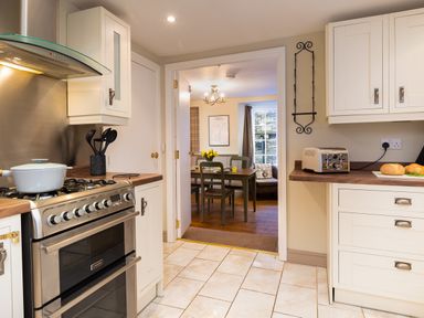 A kitchen with a stove a pot and kitchen utensils and a counter with bread leading to a dining area with a table and chairs at Ghyll Foot Cottage in Ambleside