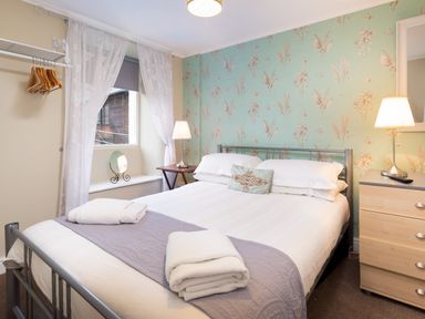 A bedroom with a bed folded towels bedside tables lamps a window with curtains and floral wallpaper at Ghyll Foot Cottage in Ambleside