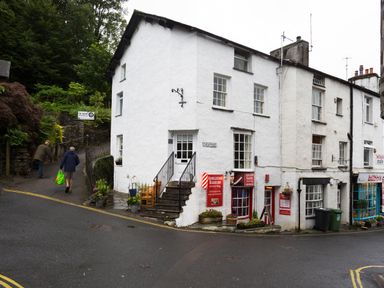 A street corner with a white building housing Ambleside Barbers and bilbos cafe with two people walking on a pathway at Ghyll Foot Cottage in Ambleside