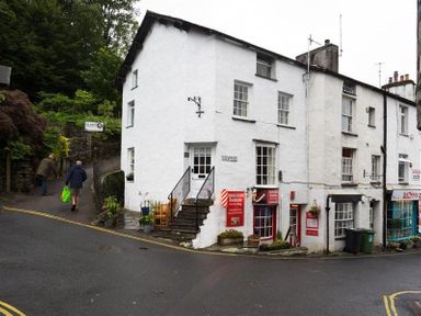 A street corner with white buildings housing Ambleside bakers and bilbo's cafe with two people walking on a path in Ambleside