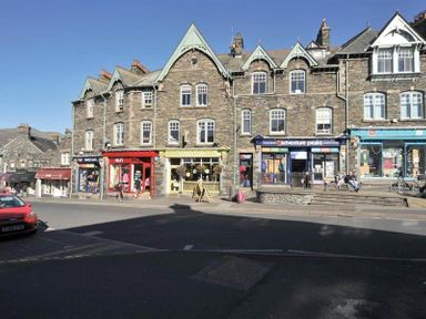 A street view with stone buildings and various shops including one with a red facade and another with a blue facade with people outside at Ghyll Foot Cottage in Ambleside