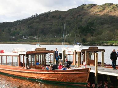 A wooden boat with people on a lake docked next to another boat with hills and trees in the background at Ghyll Foot Cottage in Ambleside
