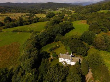 An aerial view of a white house surrounded by trees and green fields at Fair Rigg Old Farm in Cartmel