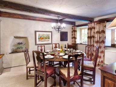 A dining room with a round table set for six with wooden chairs and patterned curtains at Fair Rigg Old Farm in Cartmel